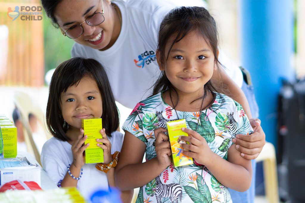 two little girls smiling while posing with their nutritional supplements and a volunteer