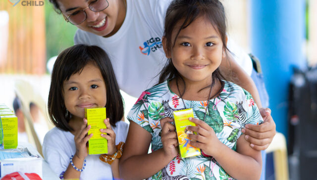 two little girls smiling while posing with their nutritional supplements and a volunteer